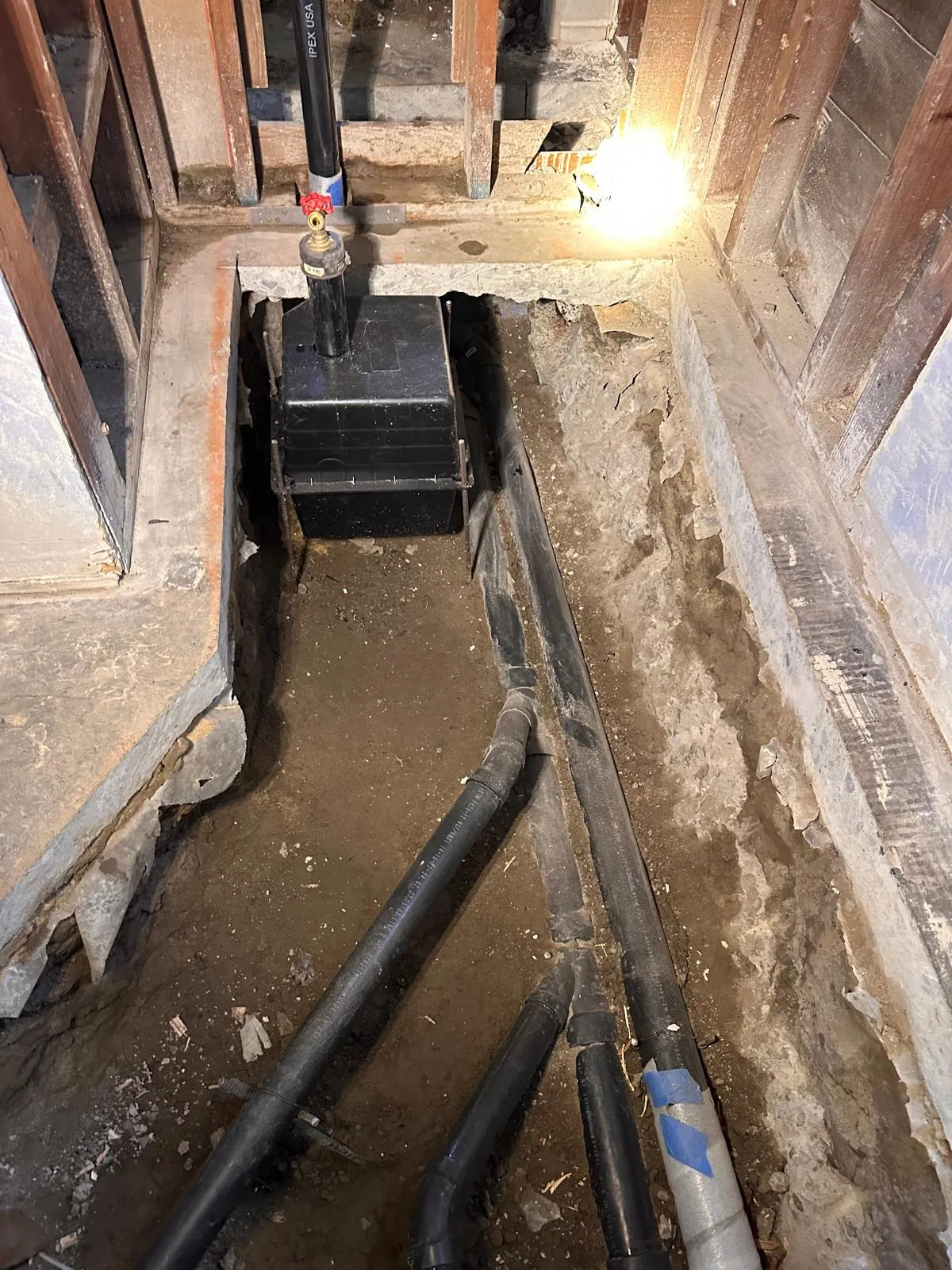 Open trench under a house slab with new drain pipe run and a work light in the background